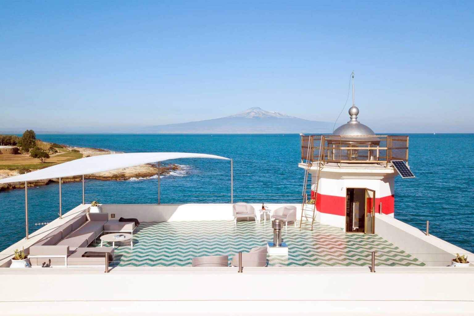 Scenic view of a modern lighthouse with a rooftop terrace overlooking the ocean, featuring lounge seating and a clear blue sky, with a distant mountain in the background. | Only on SopranoVillas