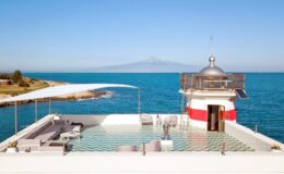 Scenic view of a modern lighthouse with a rooftop terrace overlooking the ocean, featuring lounge seating and a clear blue sky, with a distant mountain in the background.