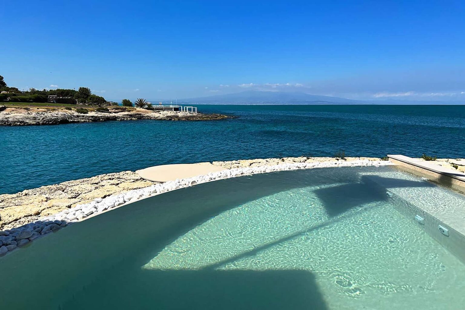 Scenic view of a tranquil coastal landscape featuring a clear swimming pool blending into the ocean, with a blue sky and distant mountains in the background. Ideal for relaxation and vacation inspiration. | Only on SopranoVillas