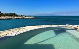 Scenic view of a tranquil coastal landscape featuring a clear swimming pool blending into the ocean, with a blue sky and distant mountains in the background. Ideal for relaxation and vacation inspiration.