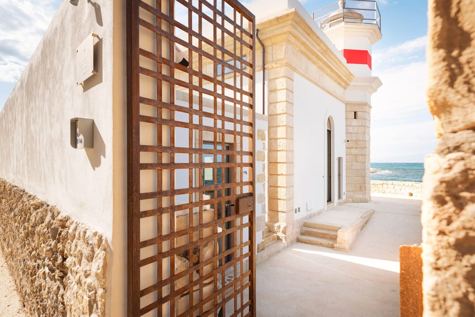Lighthouse entrance with a decorative metal gate, surrounded by stone walls and overlooking the sea under a clear blue sky. | Only on SopranoVillas