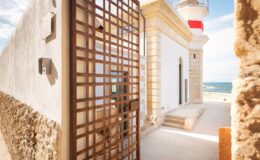 Lighthouse entrance with a decorative metal gate, surrounded by stone walls and overlooking the sea under a clear blue sky.