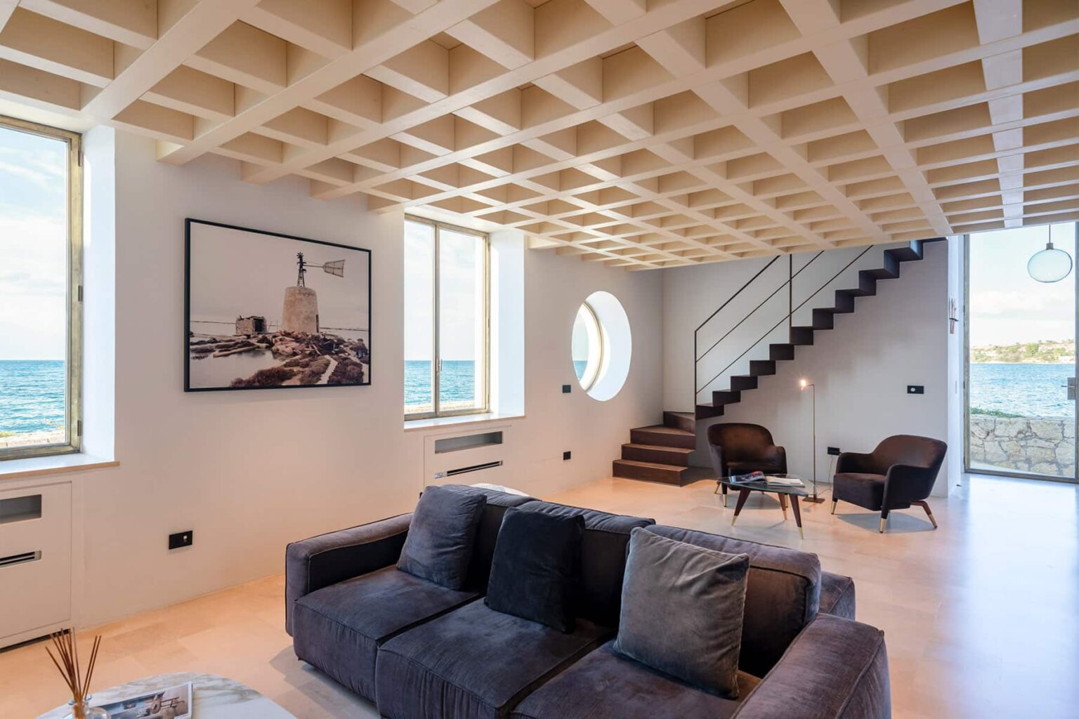 Modern coastal living room with large windows showcasing ocean views, featuring a comfortable dark sofa, minimalist decor, and a stylish staircase. | Only on SopranoVillas