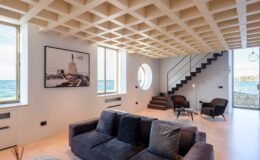 Modern coastal living room with large windows showcasing ocean views, featuring a comfortable dark sofa, minimalist decor, and a stylish staircase.