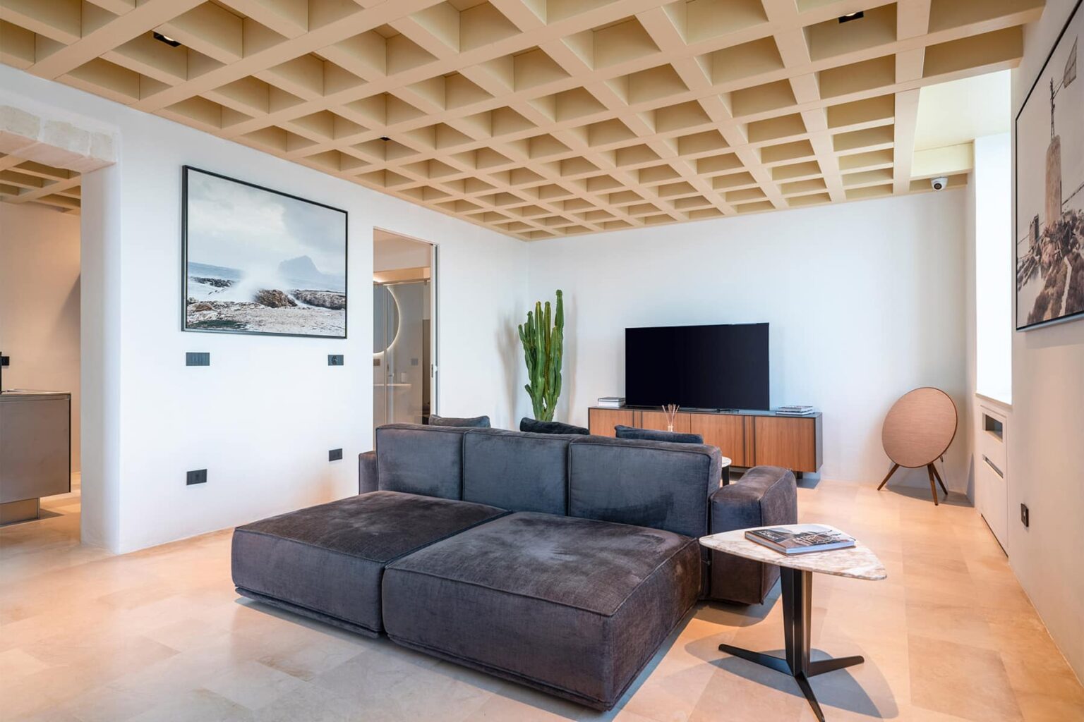 Modern living room featuring a sectional sofa, large flat-screen TV, and stylish decor. The space is illuminated by natural light and showcases a unique coffered ceiling design. | Only on SopranoVillas