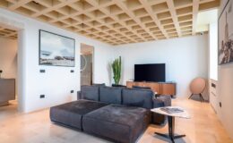 Modern living room featuring a sectional sofa, large flat-screen TV, and stylish decor. The space is illuminated by natural light and showcases a unique coffered ceiling design.