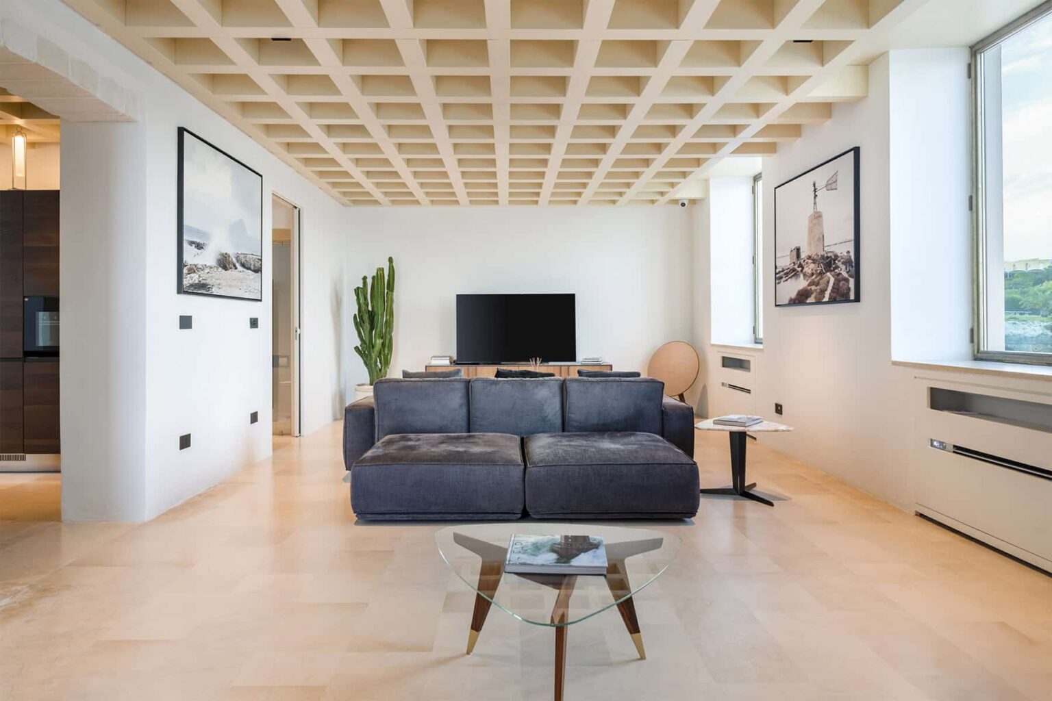 Modern living room with a dark blue sectional sofa, minimalist decor, large windows, and a stylish coffee table. The space features light wood flooring and contemporary artwork on the walls, creating a bright and inviting atmosphere. | Only on SopranoVillas