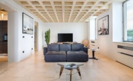 Modern living room with a dark blue sectional sofa, minimalist decor, large windows, and a stylish coffee table. The space features light wood flooring and contemporary artwork on the walls, creating a bright and inviting atmosphere.