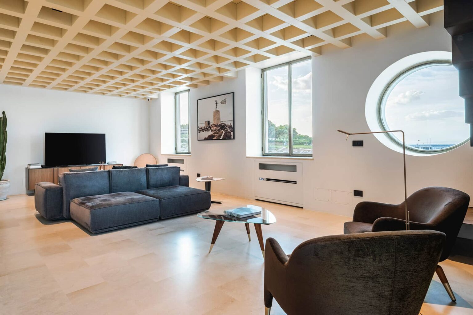 Modern living room featuring a stylish gray sectional sofa, a round coffee table, and a cozy brown armchair. Large windows provide natural light and a view of the outdoors, while the unique coffered ceiling adds architectural interest. | Only on SopranoVillas