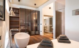 Modern bathroom interior featuring a freestanding bathtub, elegant shelving, and stylish towels neatly arranged on a countertop, with glass partitions and contemporary lighting.