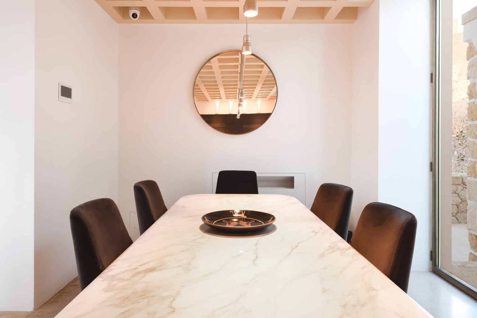 Modern dining room featuring a marble table, black leather chairs, and a round mirror. The space is illuminated by a stylish pendant light, with minimalist decor and neutral walls, creating an inviting atmosphere. | Only on SopranoVillas