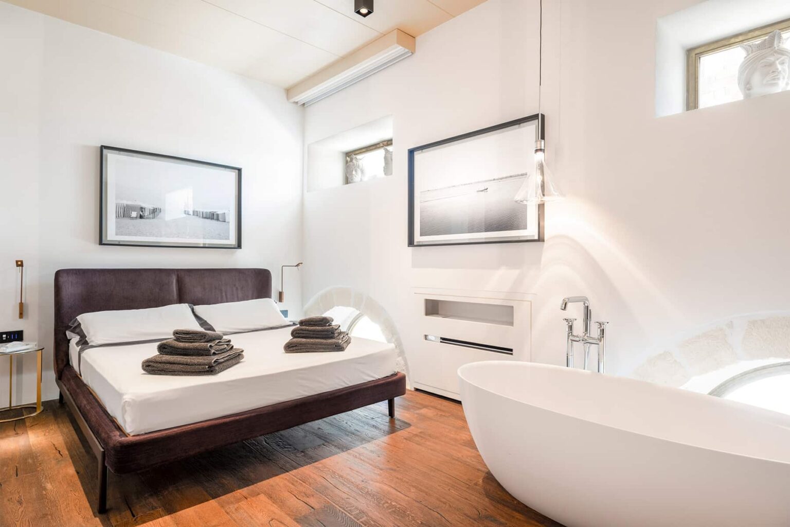 Modern bedroom with a wooden floor, featuring a double bed with neatly arranged towels, a freestanding bathtub, and large windows allowing natural light. Stylish black and white artwork adorns the walls, enhancing the contemporary decor. | Only on SopranoVillas