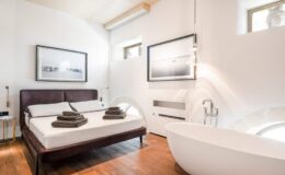 Modern bedroom with a wooden floor, featuring a double bed with neatly arranged towels, a freestanding bathtub, and large windows allowing natural light. Stylish black and white artwork adorns the walls, enhancing the contemporary decor.