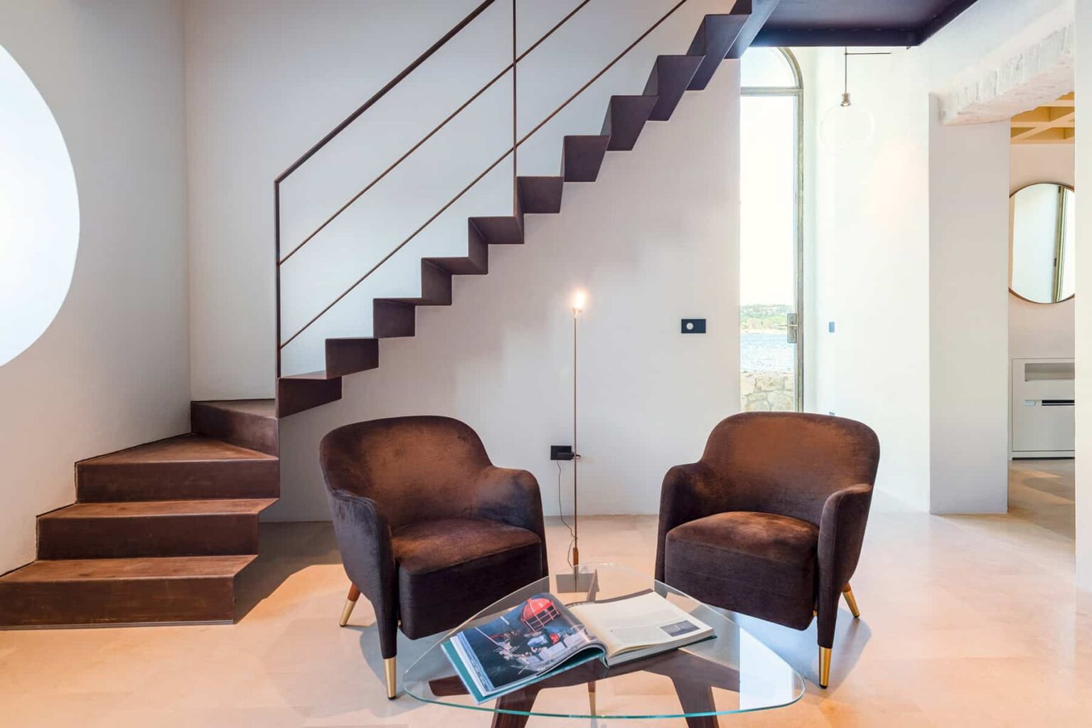 Modern interior design featuring a sleek staircase, two comfortable chairs, and a glass coffee table. The space is brightened by natural light and showcases minimalist decor with a contemporary aesthetic. | Only on SopranoVillas