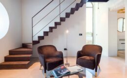 Modern interior design featuring a sleek staircase, two comfortable chairs, and a glass coffee table. The space is brightened by natural light and showcases minimalist decor with a contemporary aesthetic.