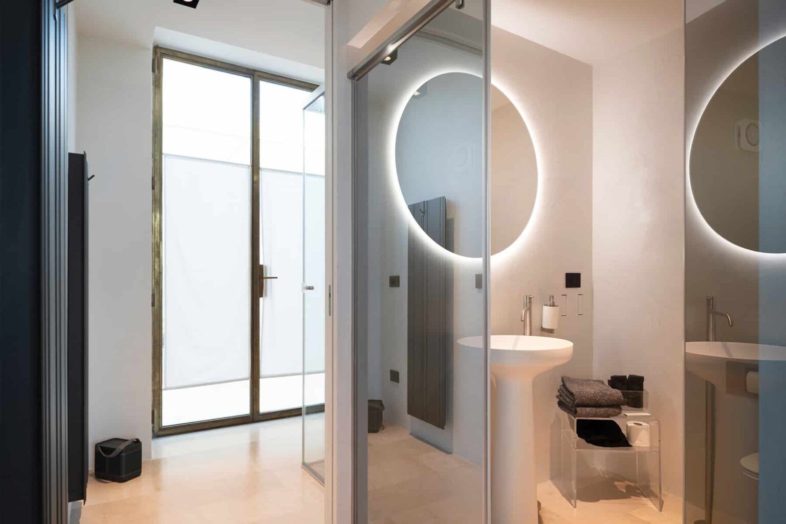 Modern bathroom interior featuring sleek glass partitions, a circular mirror with integrated lighting, and minimalist design elements. Natural light filters through a large window, enhancing the spacious feel. | Only on SopranoVillas