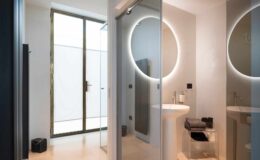 Modern bathroom interior featuring sleek glass partitions, a circular mirror with integrated lighting, and minimalist design elements. Natural light filters through a large window, enhancing the spacious feel.