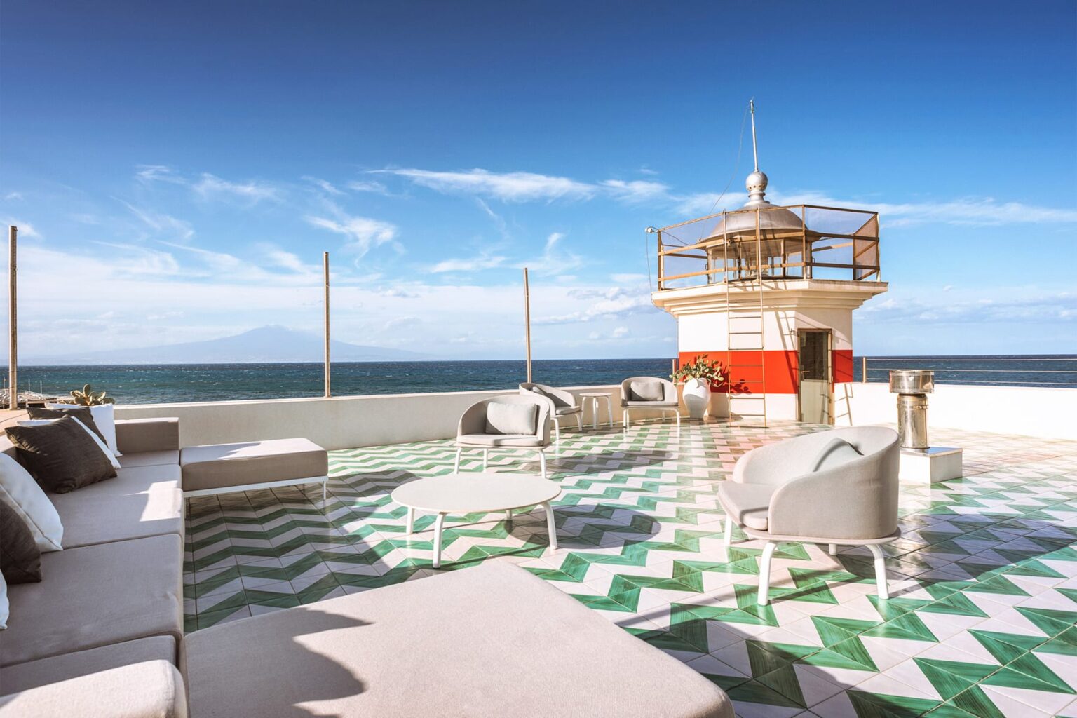 Rooftop terrace with modern seating, featuring a lighthouse and ocean view under a clear blue sky. The floor is designed with green and white geometric patterns, creating a stylish outdoor lounge area. | Only on SopranoVillas