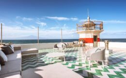 Rooftop terrace with modern seating, featuring a lighthouse and ocean view under a clear blue sky. The floor is designed with green and white geometric patterns, creating a stylish outdoor lounge area.