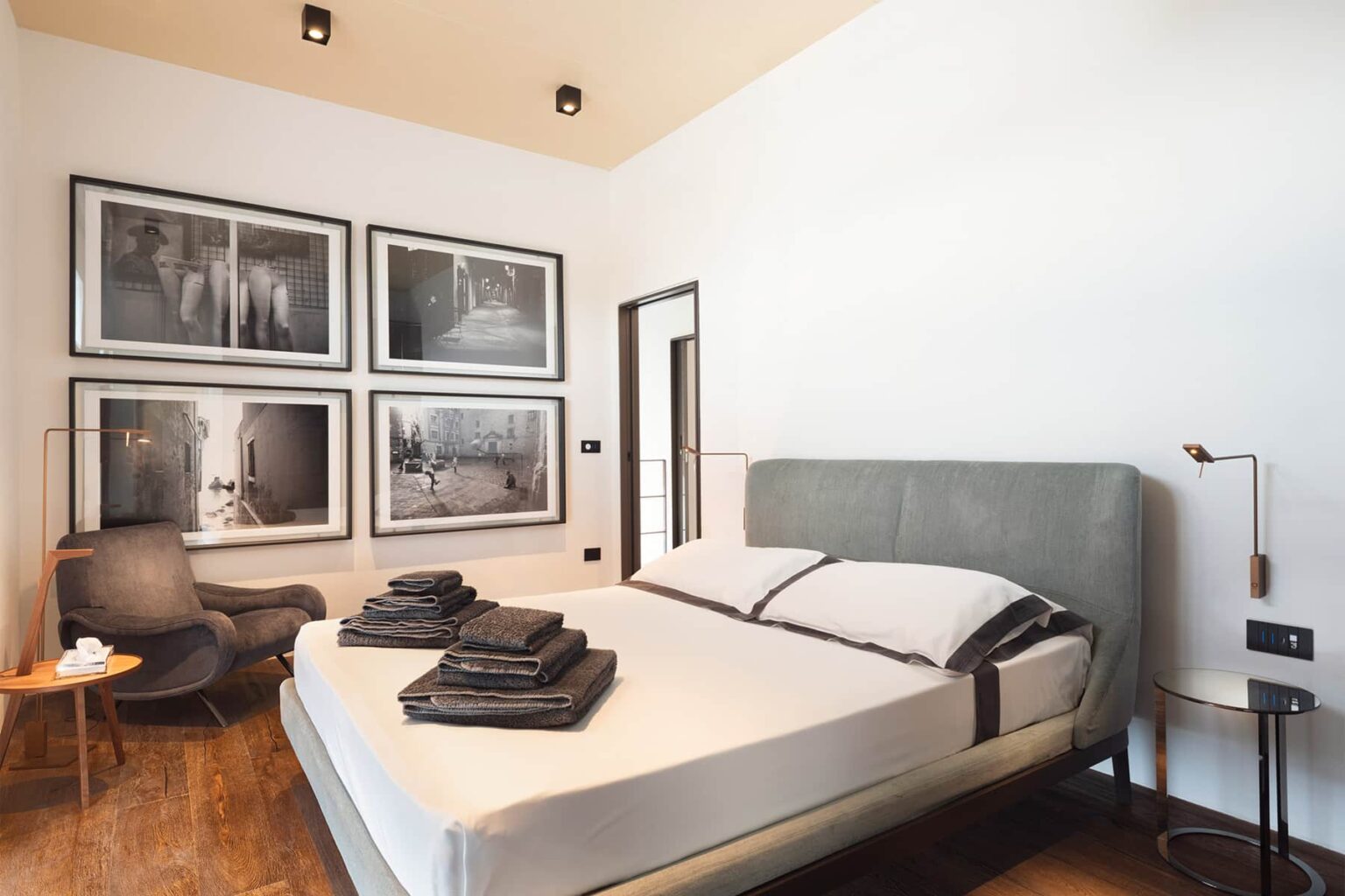 Modern bedroom featuring a neatly made bed with gray linens, a stylish armchair, and framed black-and-white photographs on the wall, showcasing a contemporary design aesthetic. | Only on SopranoVillas