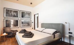 Modern bedroom featuring a neatly made bed with gray linens, a stylish armchair, and framed black-and-white photographs on the wall, showcasing a contemporary design aesthetic.