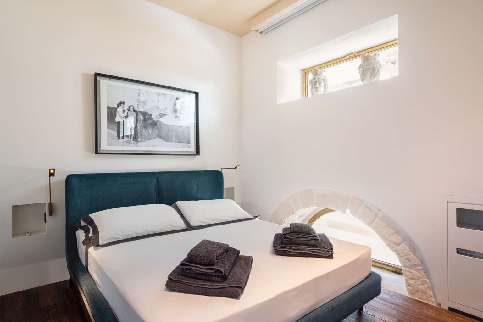 Modern bedroom featuring a comfortable bed with neatly arranged towels, minimalist decor, and a large artwork on the wall, illuminated by natural light from a window. | Only on SopranoVillas