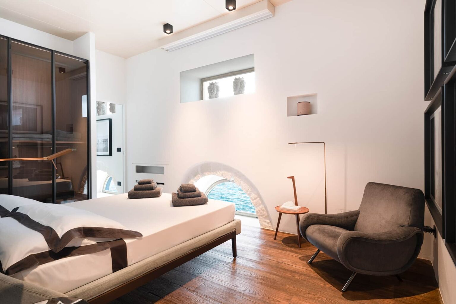 Modern bedroom with minimalist design featuring a bed with gray linens, a cozy armchair, wooden flooring, and large windows allowing natural light. | Only on SopranoVillas