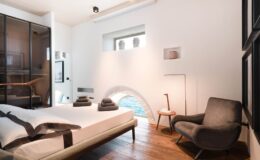 Modern bedroom with minimalist design featuring a bed with gray linens, a cozy armchair, wooden flooring, and large windows allowing natural light.
