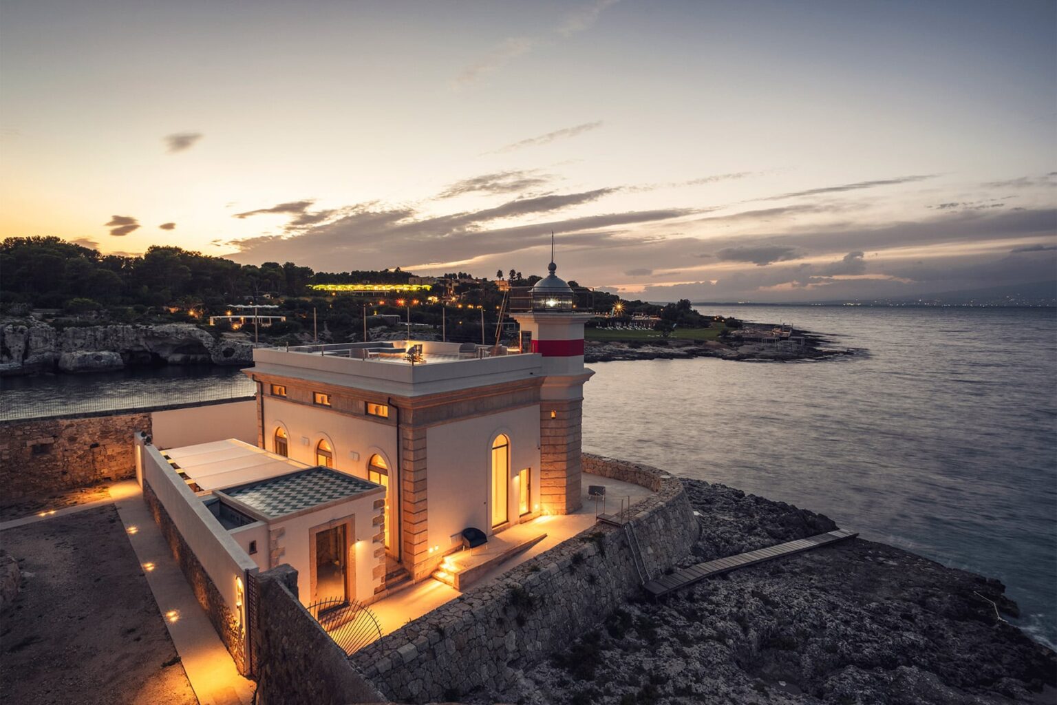 Scenic view of a historic lighthouse at sunset, illuminated against the backdrop of the ocean and rocky coastline, highlighting its architectural features and tranquil surroundings. | Only on SopranoVillas