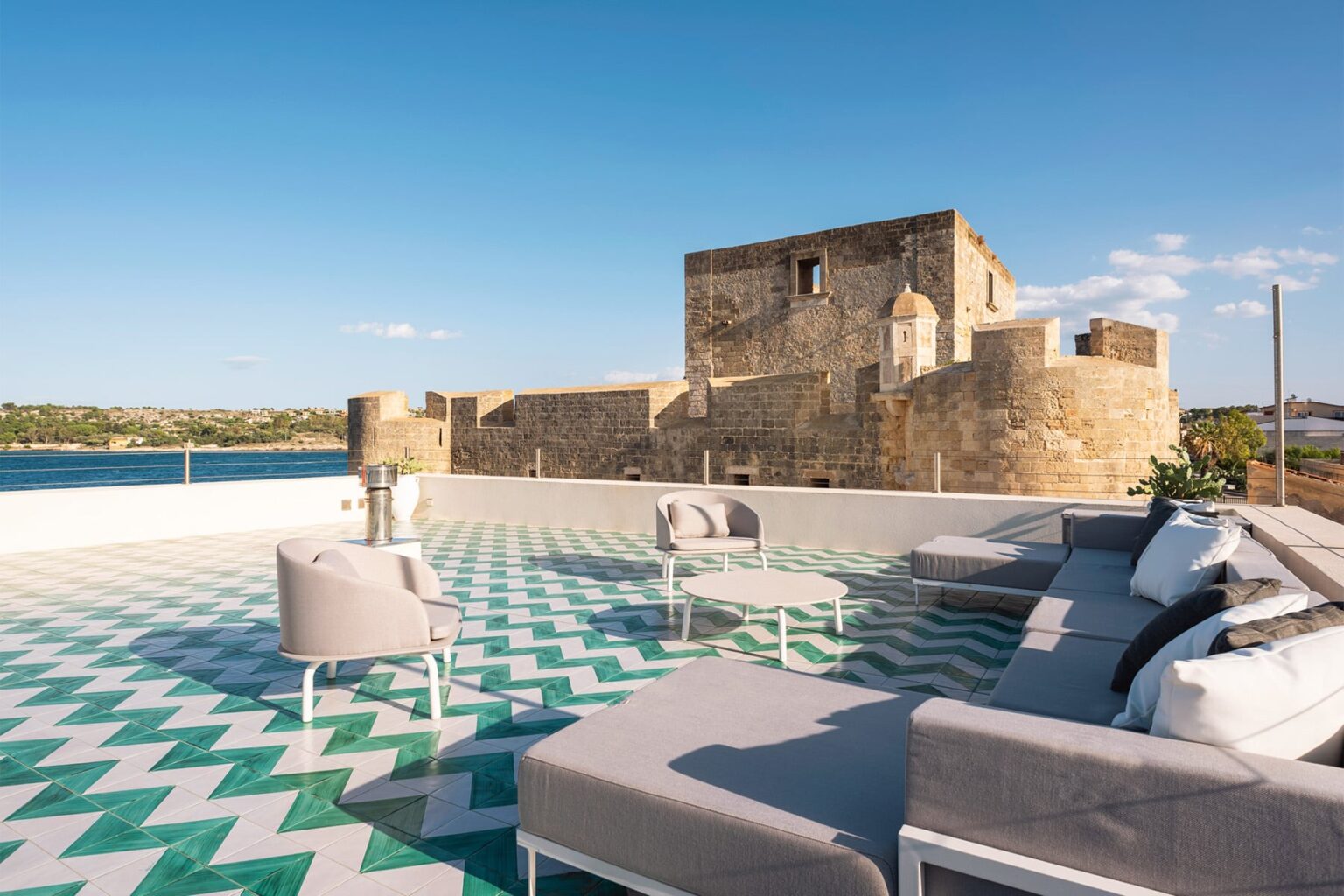 Stylish outdoor terrace with modern furniture overlooking a historic stone castle and tranquil water, featuring a geometric patterned floor. | Only on SopranoVillas