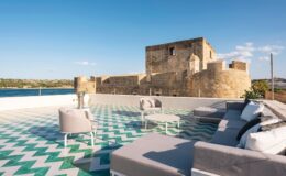 Stylish outdoor terrace with modern furniture overlooking a historic stone castle and tranquil water, featuring a geometric patterned floor.