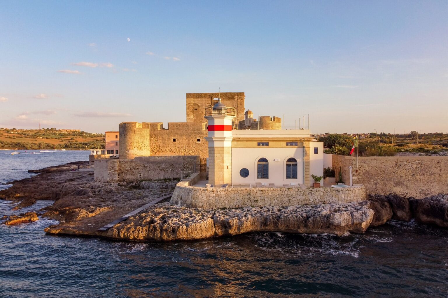 Historic coastal fortification with a lighthouse at sunset, surrounded by rocky shoreline and calm waters. | Only on SopranoVillas