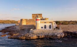 Historic coastal fortification with a lighthouse at sunset, surrounded by rocky shoreline and calm waters.