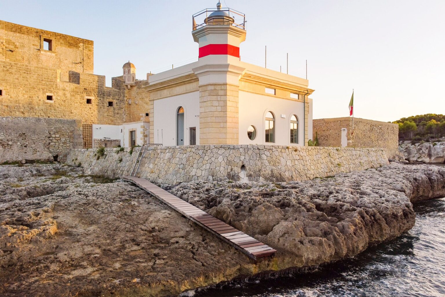 Historic lighthouse on rocky coastline, featuring a white and red structure with a walkway leading to the water, surrounded by ancient stone walls and greenery in the background. Ideal for coastal photography and maritime heritage. | Only on SopranoVillas