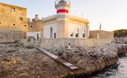Historic lighthouse on rocky coastline, featuring a white and red structure with a walkway leading to the water, surrounded by ancient stone walls and greenery in the background. Ideal for coastal photography and maritime heritage.