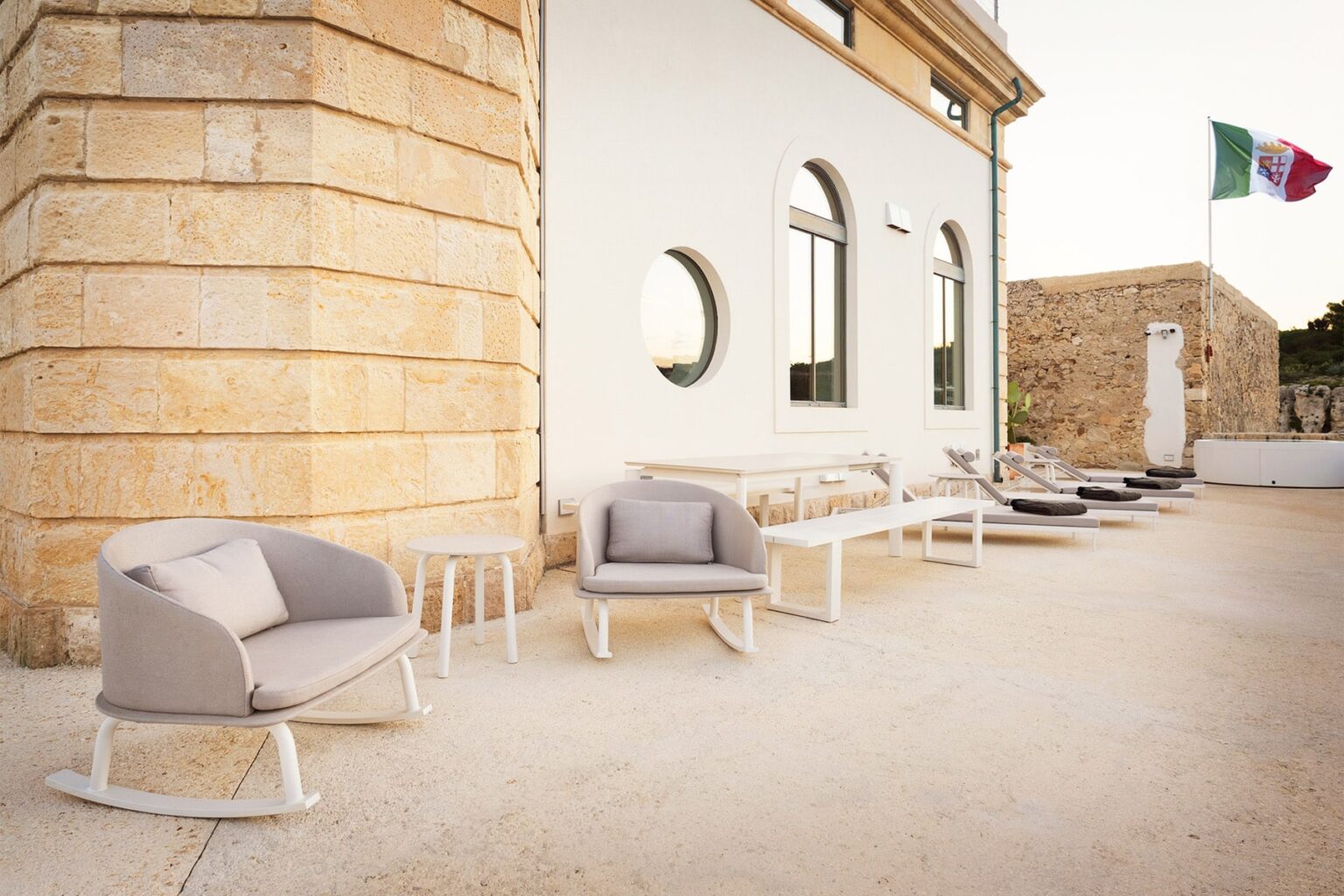 Modern outdoor seating area featuring stylish gray armchairs and tables beside a white stone building, with an Italian flag in the background. Ideal for relaxation and social gatherings in a scenic setting. | Only on SopranoVillas