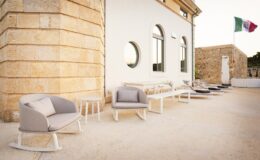 Modern outdoor seating area featuring stylish gray armchairs and tables beside a white stone building, with an Italian flag in the background. Ideal for relaxation and social gatherings in a scenic setting.