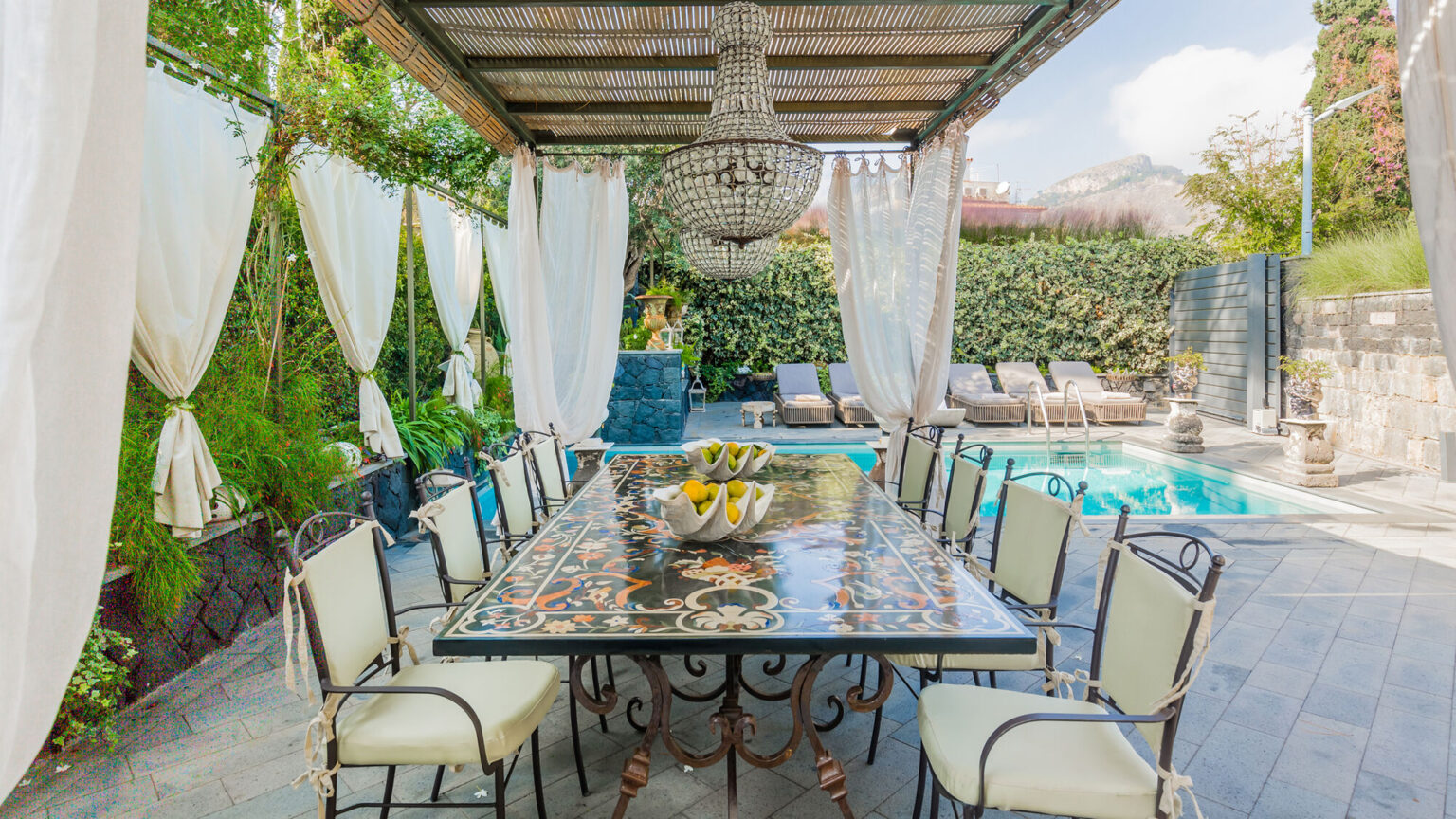 Outdoor dining area featuring a stylish table with decorative tile work, surrounded by elegant chairs and draped curtains, adjacent to a sparkling pool and lush greenery. | Only on SopranoVillas