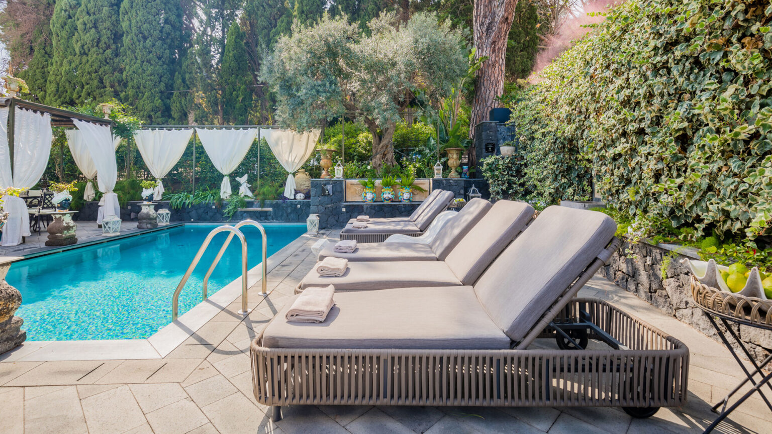 Luxurious outdoor pool area featuring comfortable lounge chairs beside a sparkling swimming pool, surrounded by lush greenery and elegant decor. Perfect for relaxation and sunbathing. | Only on SopranoVillas