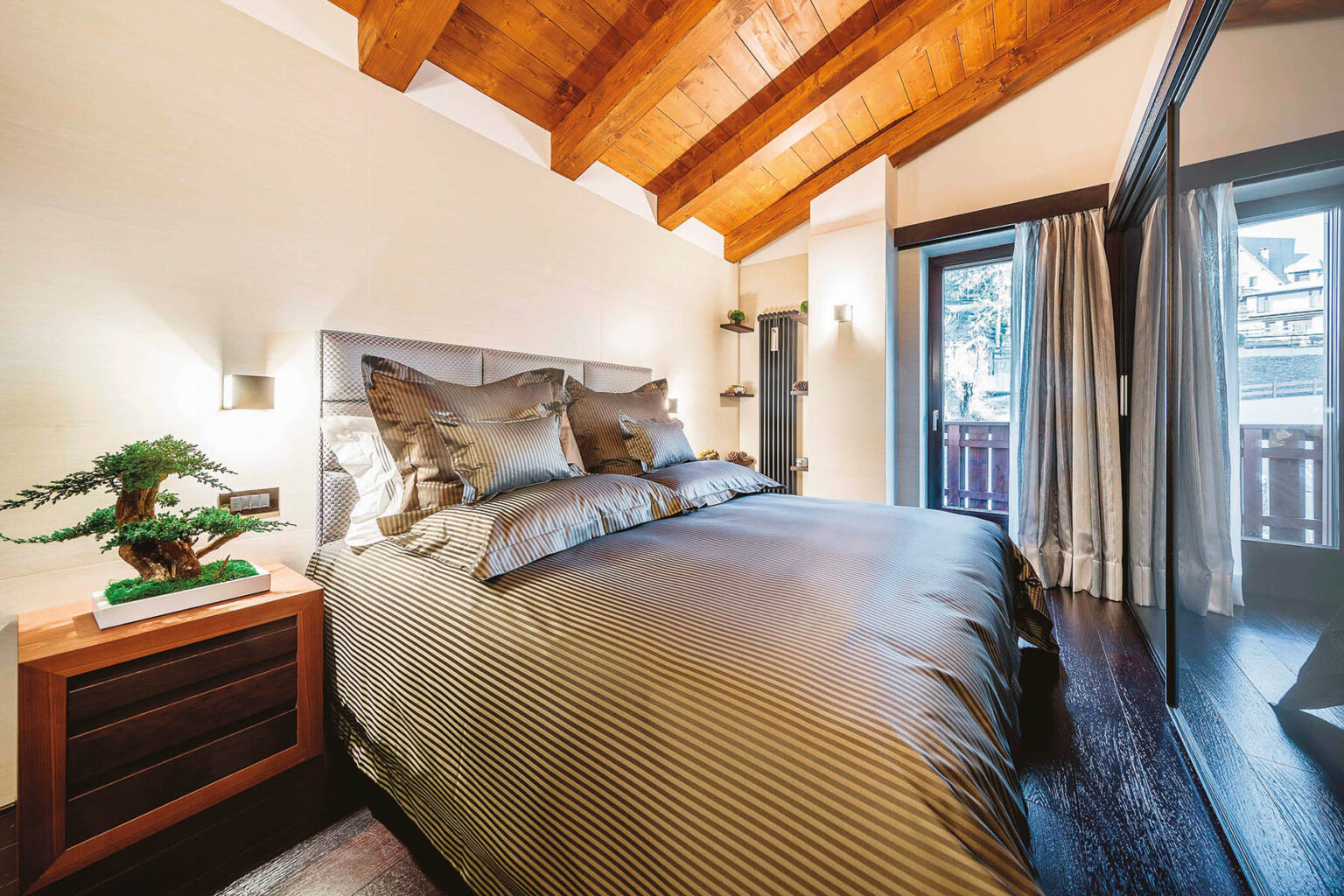 Cozy bedroom featuring a king-sized bed with elegant bedding, wooden beams, and large windows that provide natural light and a view of the outdoors. The room includes a stylish nightstand with a decorative plant. | Only on SopranoVillas