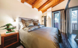Cozy bedroom featuring a king-sized bed with elegant bedding, wooden beams, and large windows that provide natural light and a view of the outdoors. The room includes a stylish nightstand with a decorative plant.