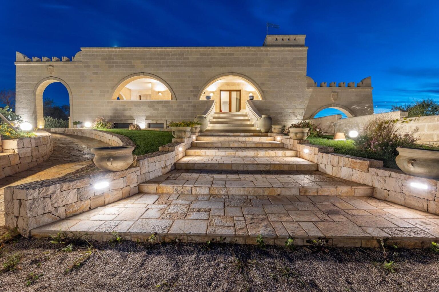 Modern stone house with illuminated pathway and landscaped garden at dusk, showcasing architectural features and inviting entrance. | Only on SopranoVillas
