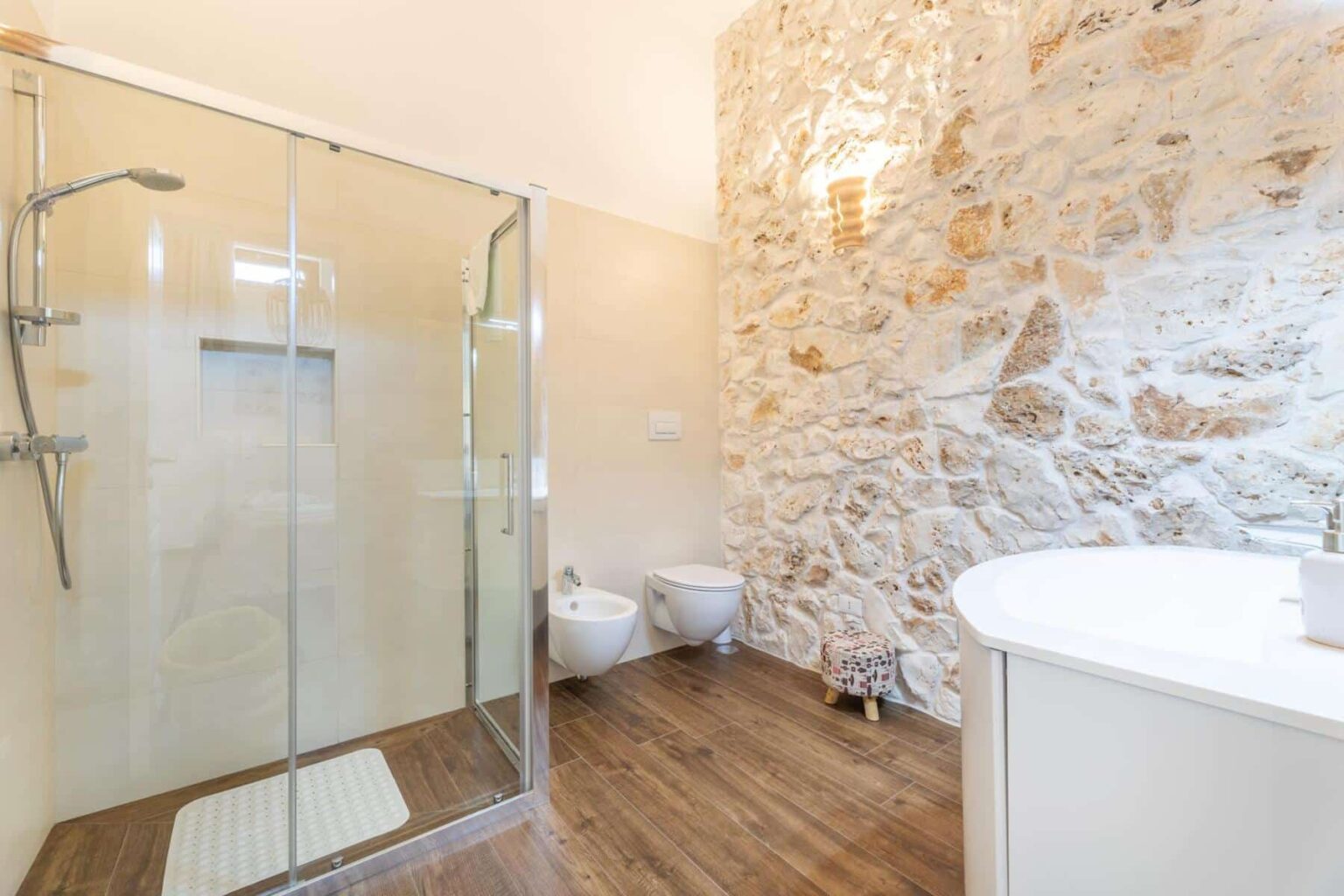 Modern bathroom featuring a glass shower enclosure, bidet, and a stylish circular sink, complemented by a rustic stone wall and wooden flooring. | Only on SopranoVillas