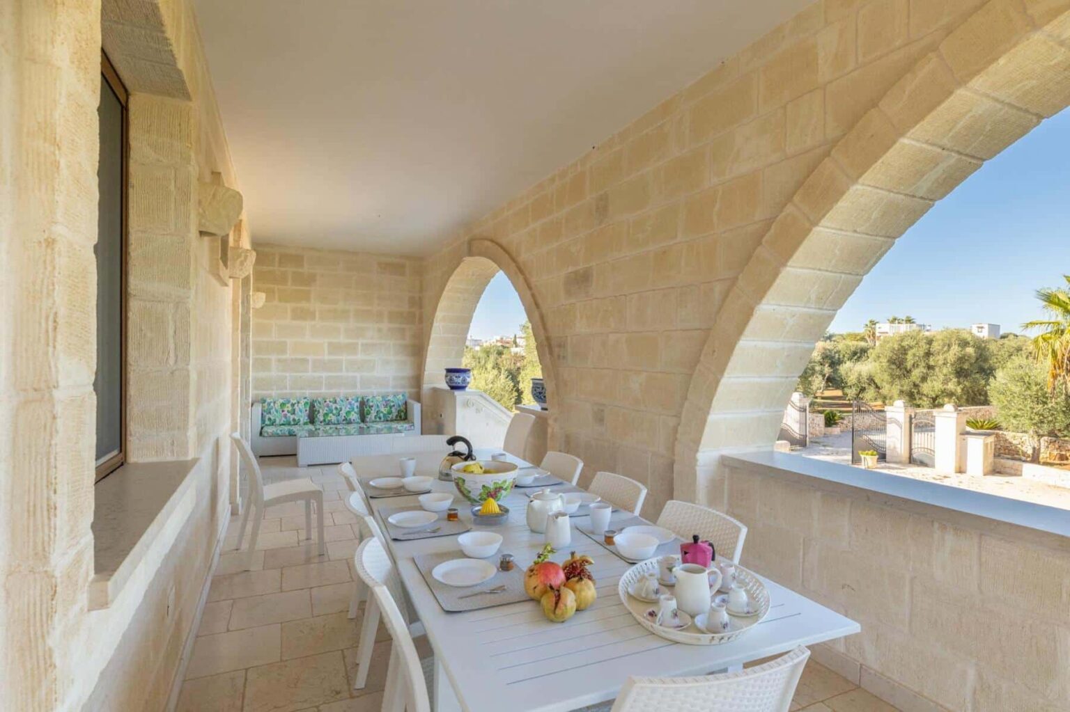 Spacious outdoor dining area featuring a white table set with dishes and fresh fruit, surrounded by stone arches and a scenic view of greenery. Ideal for alfresco dining and entertaining. | Only on SopranoVillas