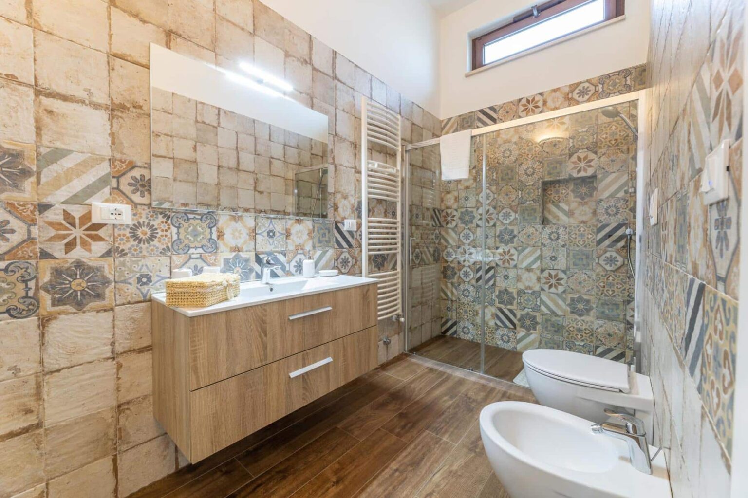 Modern bathroom featuring stylish patterned tiles, a wooden vanity with dual sinks, a large mirror, and a glass shower enclosure, complemented by a warm wood floor. | Only on SopranoVillas