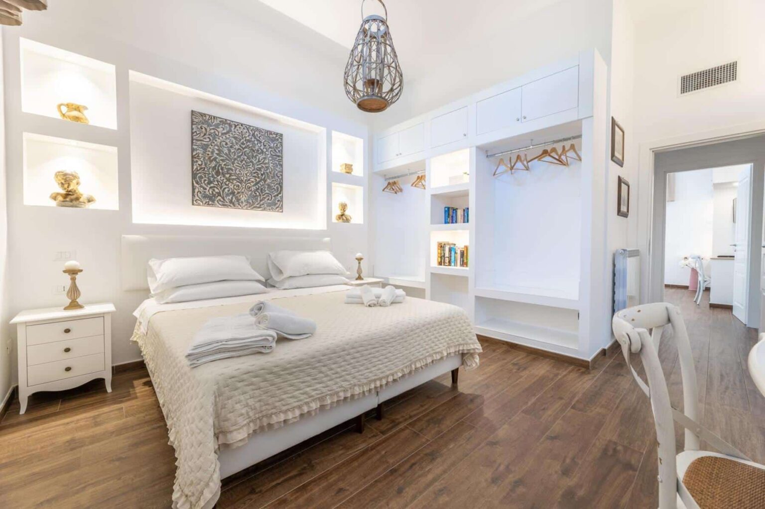 Modern bedroom interior featuring a cozy double bed with white linens, decorative wall shelves, and wooden flooring. The room is illuminated by a stylish pendant light and includes a minimalist design with a comfortable seating area and an organized closet space. | Only on SopranoVillas