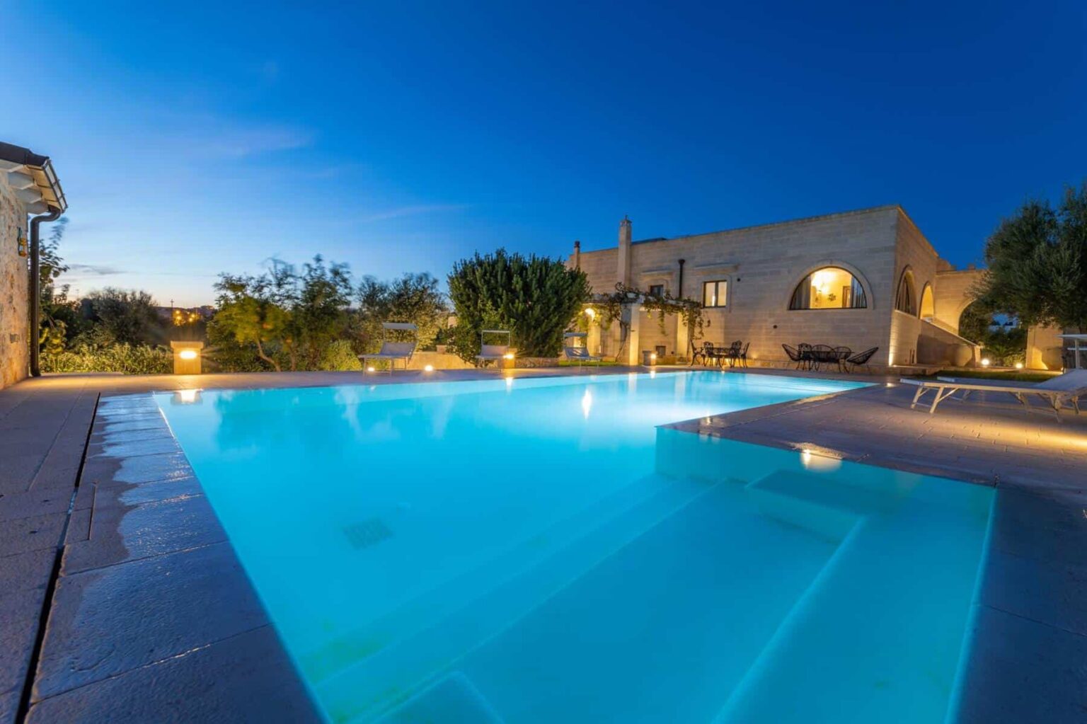 Luxurious outdoor swimming pool with illuminated edges, surrounded by lush greenery, at twilight in a modern villa setting. | Only on SopranoVillas