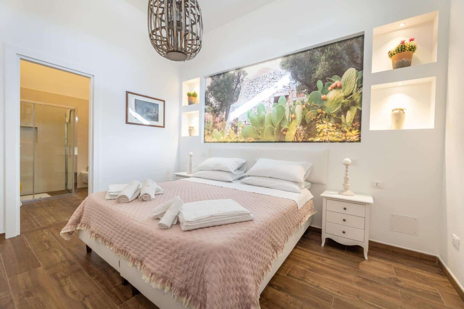 Cozy bedroom featuring a comfortable bed with a light pink quilt, neatly arranged towels, and a decorative wall art of a garden. The room includes a stylish pendant light, a nightstand with drawers, and built-in shelves displaying potted plants. A glass shower is visible in the background, highlighting the room's modern design and inviting atmosphere. | Only on SopranoVillas