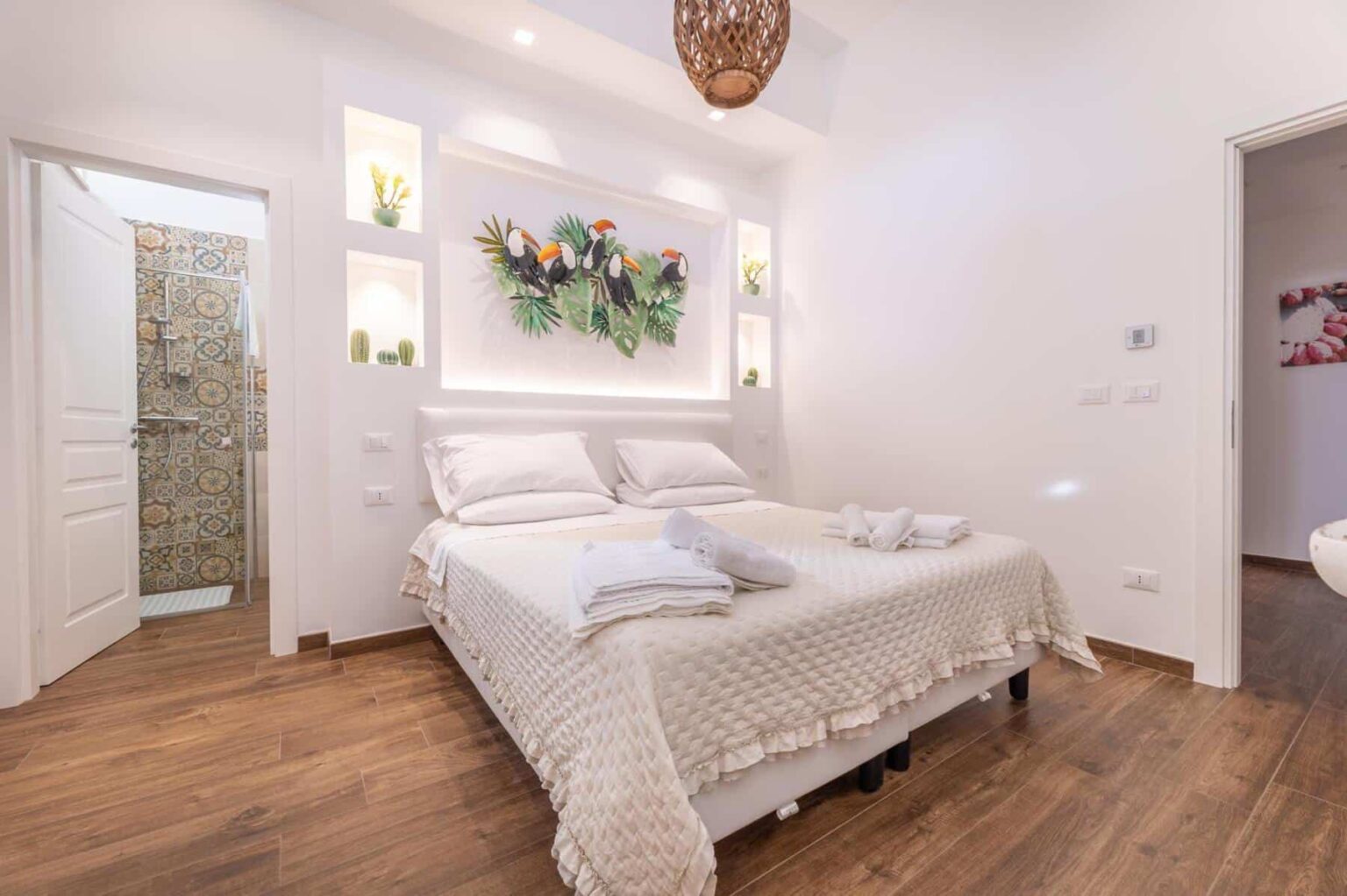 Cozy modern bedroom featuring a comfortable double bed with soft linens, decorative pillows, and a stylish wall art display of flowers. The room has warm wooden flooring and bright lighting, creating an inviting atmosphere. A doorway leads to a bathroom, enhancing the overall functionality. | Only on SopranoVillas