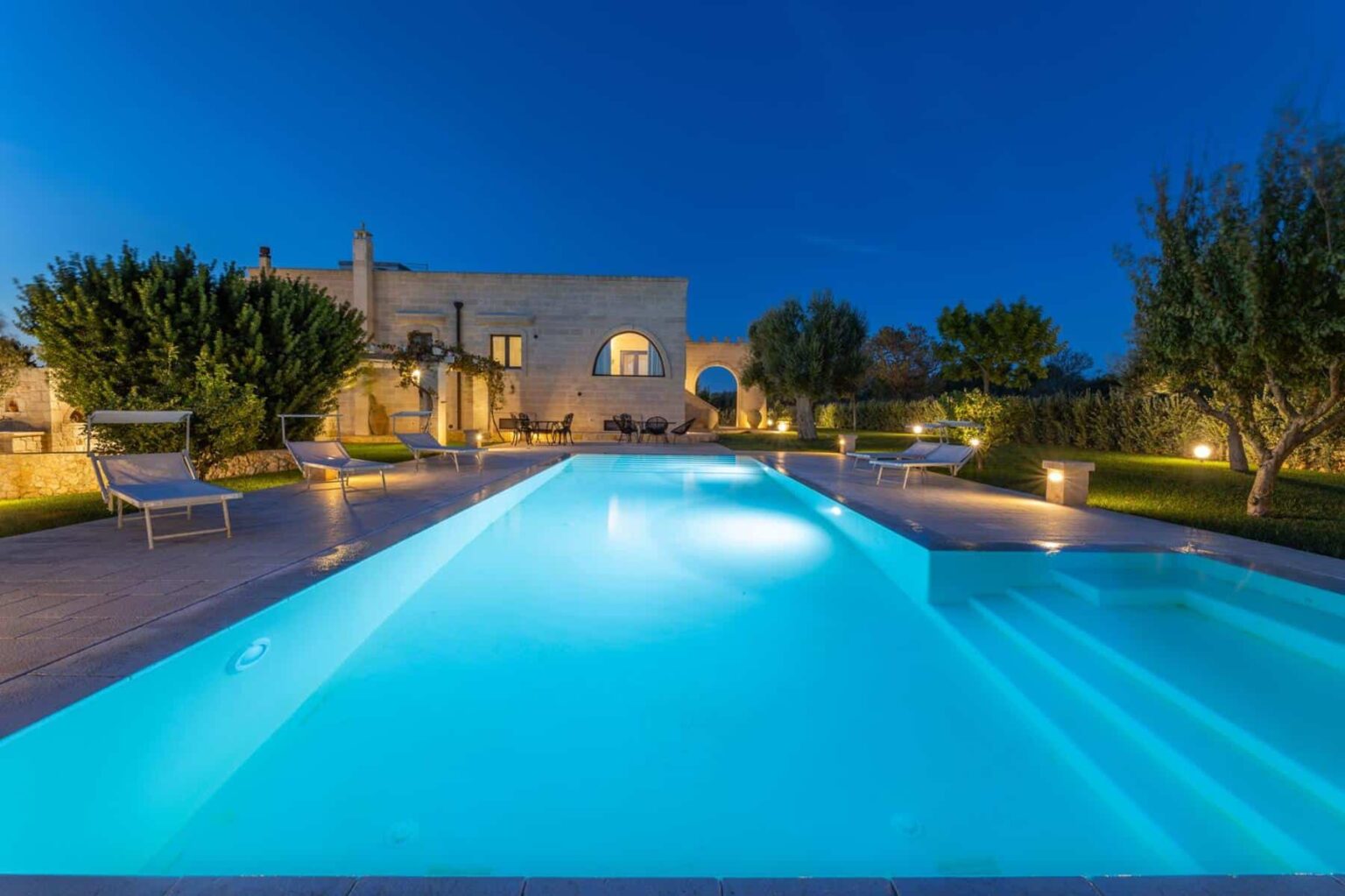 Luxury villa with a serene swimming pool illuminated at night, surrounded by lush greenery and elegant landscaping. | Only on SopranoVillas